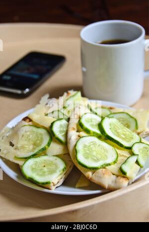 Spuntino di pane piatto scandinavo, con fette di formaggio svedese e cetrioli sottili, cosparso di sale alle erbe, con una tazza di caffè nero e iphone o Foto Stock