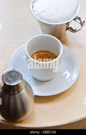 Tazza bianca in porcellana di caffè espresso appena preparato con la scritta "creme" con latte in schiuma nella brocca e shaker al cacao - in procinto di trasformare l'espresso in capp Foto Stock
