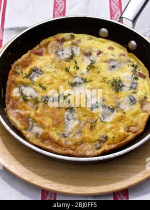 Frittata con formaggio di capra, funghi e timo, servita in padella su piatto di legno. - Foto Stock