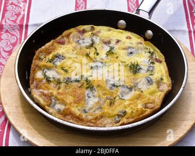 Frittata con formaggio di capra, funghi e timo, servita in padella su piatto di legno. - Foto Stock