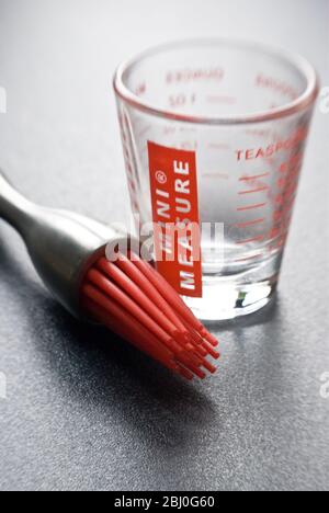 Spazzola per pasticceria moderna con setole in silicone rosso con un piccolo bicchiere graduato su superficie ruvida scura - Foto Stock