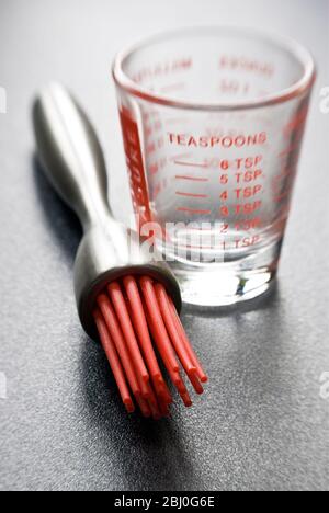 Spazzola per pasticceria moderna con setole in silicone rosso con un piccolo bicchiere graduato su superficie ruvida scura - Foto Stock
