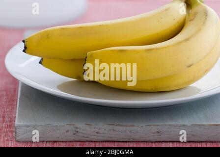 Mazzo di banane su piatto bianco, su tavola grigia, con fondo rosso - Foto Stock