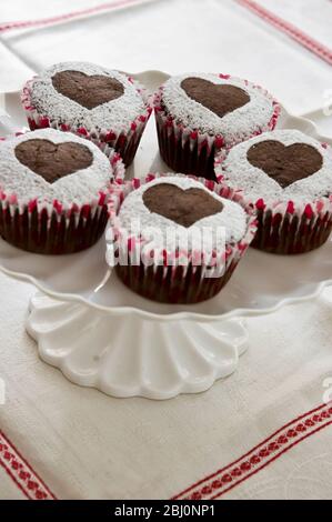 Cinque muffin al cioccolato decorati con zucchero a velo a forma di cuore su base per torta bianca - Foto Stock