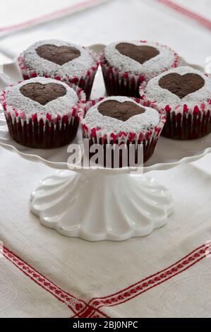 Cinque muffin al cioccolato decorati con zucchero a velo a forma di cuore su base per torta bianca - Foto Stock