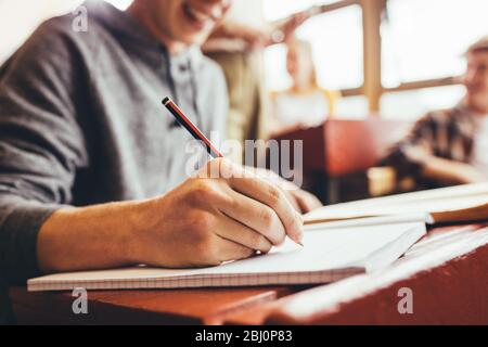 Primo piano di studenti maschi che prendono appunti seduti alla scrivania durante la lezione nella scuola superiore. Concentrarsi sulla scrittura a mano in notebook con una matita. Foto Stock