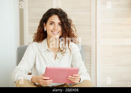 Donna caucasica attraente con capelli ricci che indossa camicia bianca tenendo computer tablet rosa guardando la fotocamera sorridente Foto Stock