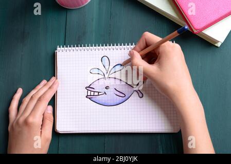 Le mani femminili disegnano balena in taccuino su sfondo tavolo di legno Foto Stock