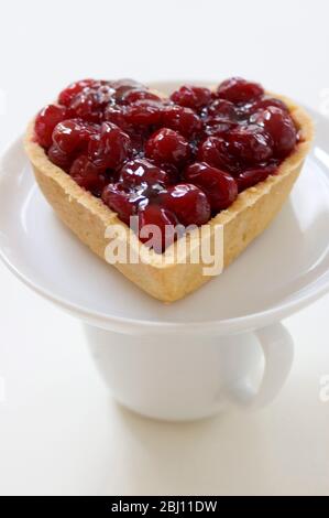 Crostata di ciliegia a forma di cuore su piattino bianco equilibrante su tazza - Foto Stock