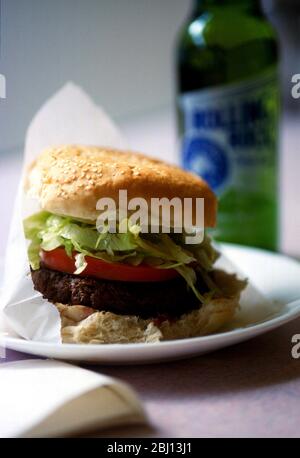 Hamburger americano classico con pomodoro e lattuga grattugiata in panino di sesamo servito in sacchetto di carta su piatto bianco con bottiglia di birra Rolling Rock, a forma Foto Stock