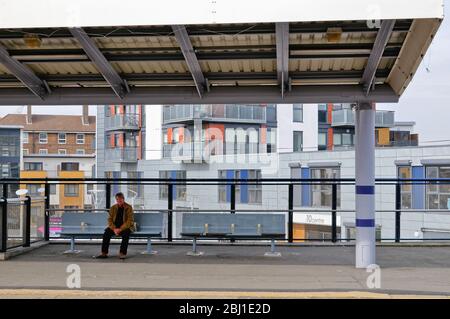 Un uomo di mezza età seduto sulla piattaforma della stazione della linea principale di Greenwich in attesa di un treno con appartamenti moderni dietro Londra Inghilterra Regno Unito Foto Stock