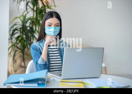 Donna dai capelli scuri seduta alla scrivania. Guardando il laptop, indossando una maschera protettiva Foto Stock