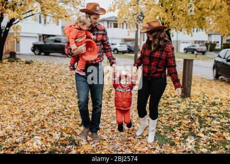 Una famiglia con due bambini vestiti da vigili del fuoco per Halloween cammina attraverso un parco di foglie sparse. Foto Stock