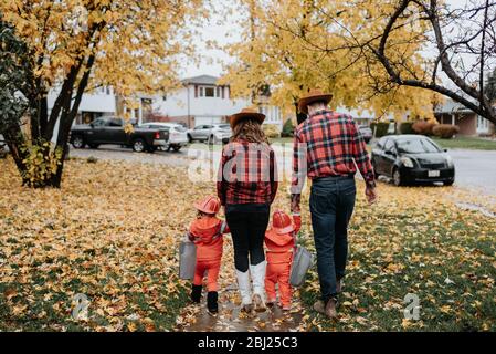 Una famiglia con due bambini vestiti da vigili del fuoco per Halloween cammina attraverso un parco di foglie sparse. Foto Stock