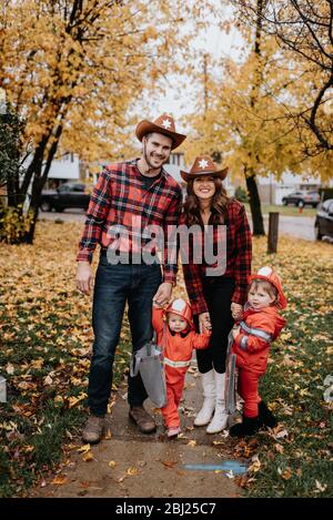 Una famiglia con due bambini vestiti da pompieri per Halloween posa per una foto in un parco di foglie sparse. Foto Stock