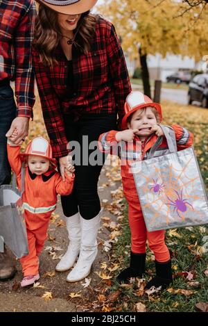 Una famiglia con due bambini vestiti da vigili del fuoco per Halloween cammina attraverso un parco di foglie sparse. Foto Stock