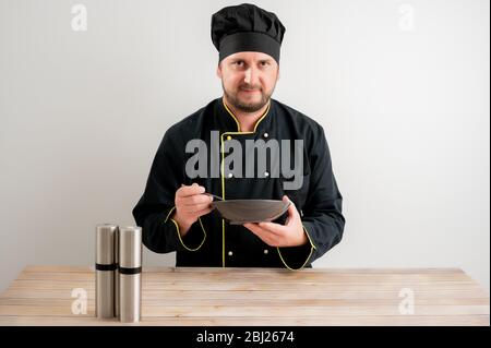 Ritratto di giovane cuoco in uniforme nera mostra il piatto in mano posando con sale su sfondo bianco isolato Foto Stock