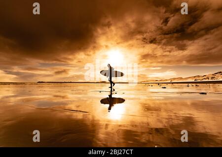 Silhouette di una donna che porta una tavola da surf che cammina attraverso una spiaggia con un tramonto sullo sfondo. Foto Stock
