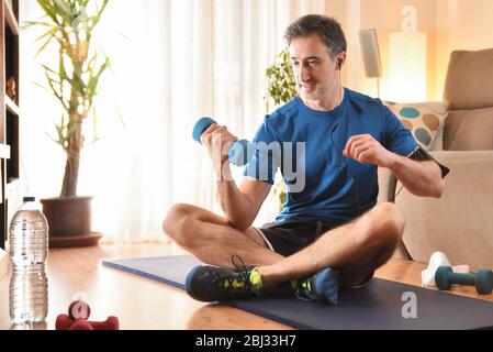L'uomo che fa il dumbbell si sport ascoltando la musica con le cuffie sul telefono seduto su un tappetino nel suo soggiorno Foto Stock