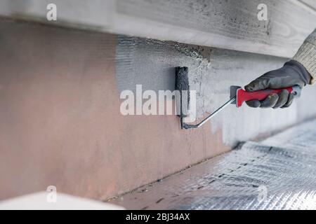 Uomo che dipinge casa socle con rullo di vernice. Primo piano della vernice grigia arrotolabile sulla base dell'edificio. Ristrutturazione domestica Foto Stock