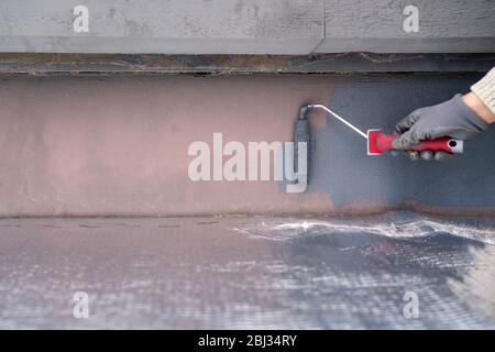 Uomo che dipinge casa socle con rullo di vernice. Primo piano della vernice grigia arrotolabile sulla base dell'edificio. Ristrutturazione domestica Foto Stock