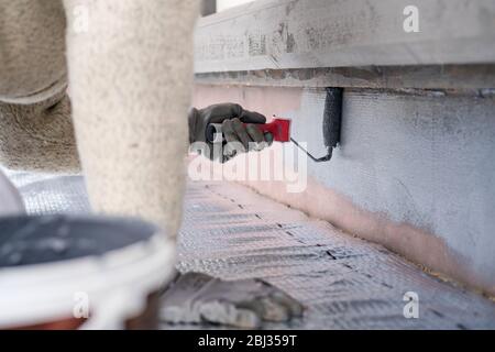 Uomo che dipinge casa socle con rullo di vernice. Primo piano della vernice grigia arrotolabile sulla base dell'edificio. Ristrutturazione domestica Foto Stock
