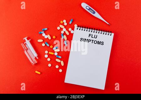 Notepad, una varietà di medicinali, termometro, siringa e fiale con medicinale su sfondo rosso. Scritto su un blocco note SINTOMI DI INFLUENZA. Foto Stock