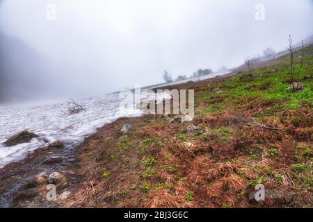 L'ultima neve d'inverno si scioglie sulla collina in alta montagna contro le nuvole scure. I resti di neve sporca davanti ad una sorgente calda. Con Foto Stock