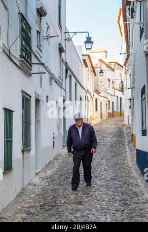 Uomo anziano in piano CAP camminare in tipico scenario di strada di case bianche e gialle, lanterne e strada stretta ciottoli a Evora, Portogallo Foto Stock