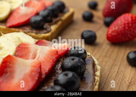 Delizioso pane tostato con guarnimento al cioccolato, banana, fragola e mirtillo su un asse di legno. Concetto di colazione gustosa e salutare. Foto Stock