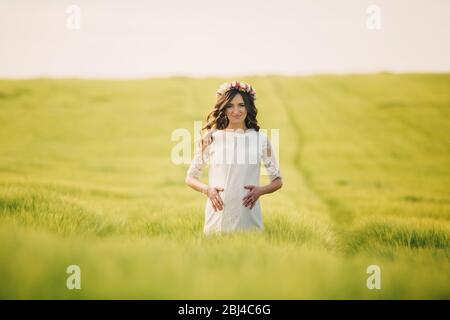 donna incinta in abito bianco con corona di fiori sulla testa sta tenendo le mani sullo stomaco e rilassandosi all'aperto in campo di erba. concetto di maternità Foto Stock