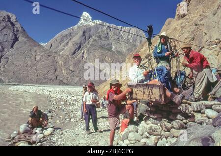 Pakistan, zone settentrionali delle montagne di Karakoram. Un gruppo europeo di trekking condivide il compito comune di caricare la scatola Jola per aiutare l'attraversamento del fiume Braldu alla testa della gola di Braldu. La Jola è stata sostituita da un ponte di sospensione che rende l'operazione più facile e sicura Foto Stock