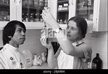 Anni '70, storico, all'interno di una stanza con scaffali di bottiglie di farmaci, una infermiera femminile, possibile un infermiere studente, mettendo il liquido da una bottiglia in una siringa, con un giovane infermiere maschio che la sovrinteista, Lewisham, Londra del Sud, Inghilterra, Regno Unito. Foto Stock