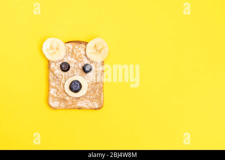 Divertente colazione per bambini brindisi con burro di arachidi a forma di orso con mirtillo e banana su sfondo giallo. Vista superiore, spazio di copia Foto Stock