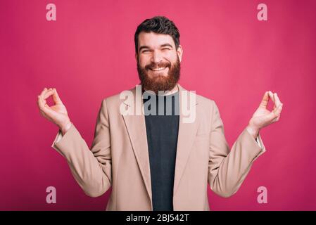 Sorridente uomo bearded divertente sta cercando di fare zen gesto con le sue mani su sfondo rosa. Foto Stock