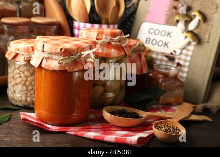 Vasetti con verdure e fagioli sottaceto, spezie, libro di ricette e utensili da cucina su sfondo legno Foto Stock