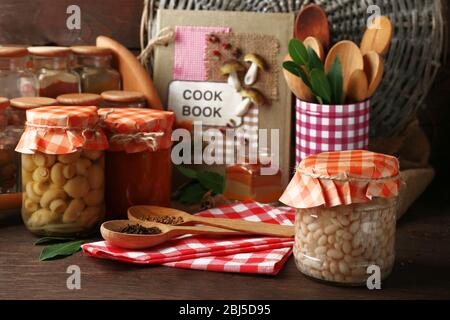 Vasetti con verdure e fagioli sottaceto, spezie, libro di ricette e utensili da cucina su sfondo legno Foto Stock