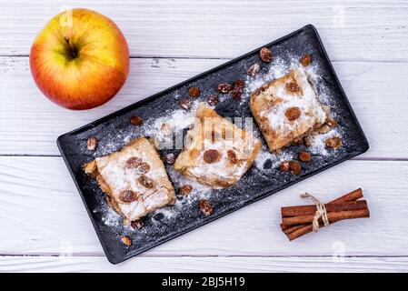 Fette di strudel di torta di mele fatta in casa in un piatto con ingredienti su un rustico tavolo di legno bianco. Torta con mele, uvetta e cannella, dolci Foto Stock