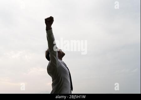 Profilo di successo del giovane uomo d'affari in piedi sotto cielo nuvoloso grigio celebrando il successo con le sue braccia sollevate in alto. Foto Stock