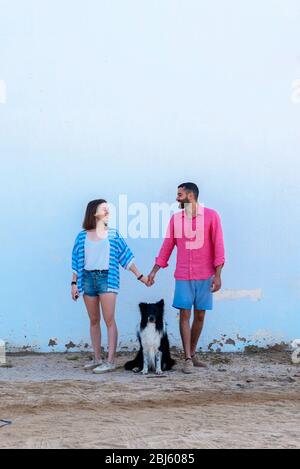 Coppia felice in piedi con cane contro il muro mentre tiene le mani Foto Stock