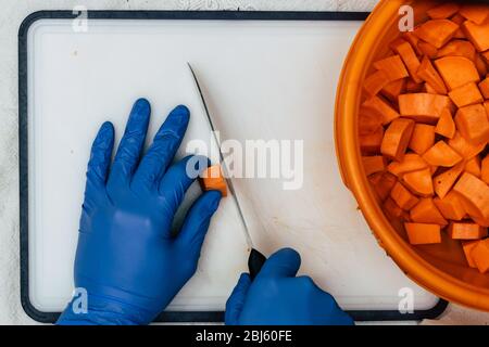 Un uomo con guanti blu taglia la patata dolce - con un coltello, quasi tagliato e pelato patate dolci in una ciotola di plastica, salvaguardie e cautela contro la corona Foto Stock