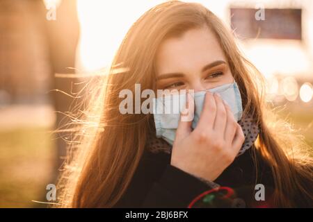 Ritratto di giovane donna felice che indossa maschera protettiva viso medico in piedi in città al tramonto Foto Stock
