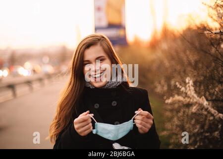 Felice giovane donna allegra rimuovere maschera medica viso mentre in piedi sulla strada in città durante il tramonto in primavera Foto Stock