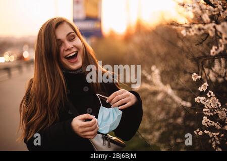 Felice giovane donna allegra rimuovere maschera medica viso mentre in piedi sulla strada in città durante il tramonto in primavera Foto Stock