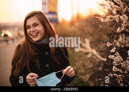 Felice giovane donna allegra rimuovere maschera medica viso mentre in piedi sulla strada in città durante il tramonto in primavera Foto Stock