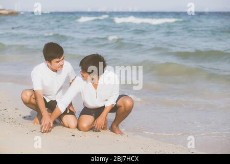 Giovane coppia asiatica gay sorridente romantico disegno forma del cuore insieme sulla sabbia in vacanza, felice omosessuale e divertente con amore seduto sulla sabbia al be Foto Stock