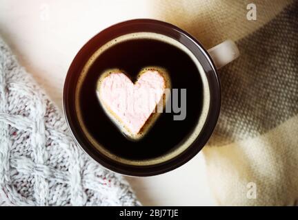 Tazza di caffè caldo con marshmallow al cuore e sciarpe calde su fondo in legno chiaro, primo piano Foto Stock
