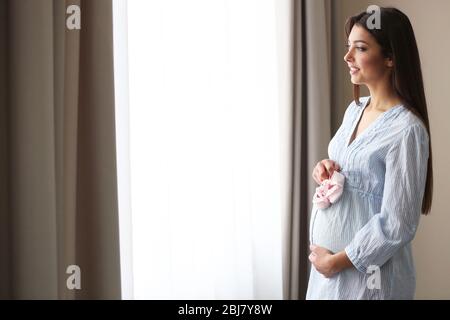 Donna incinta in piedi vicino alla finestra con il bambino bootees in camera Foto Stock