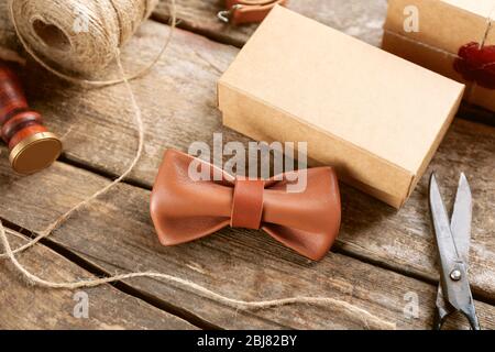 Cravatta ad arco in pelle marrone chiaro e scatola regalo in cartone su tavolo in legno, primo piano Foto Stock