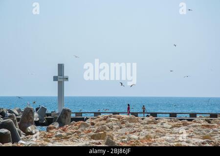 Mare, vista di una grande croce di cemento sulla baia, persone a piedi e uccelli volare, acque blu e cielo, frangiflutti, stagione estiva, natura Foto Stock